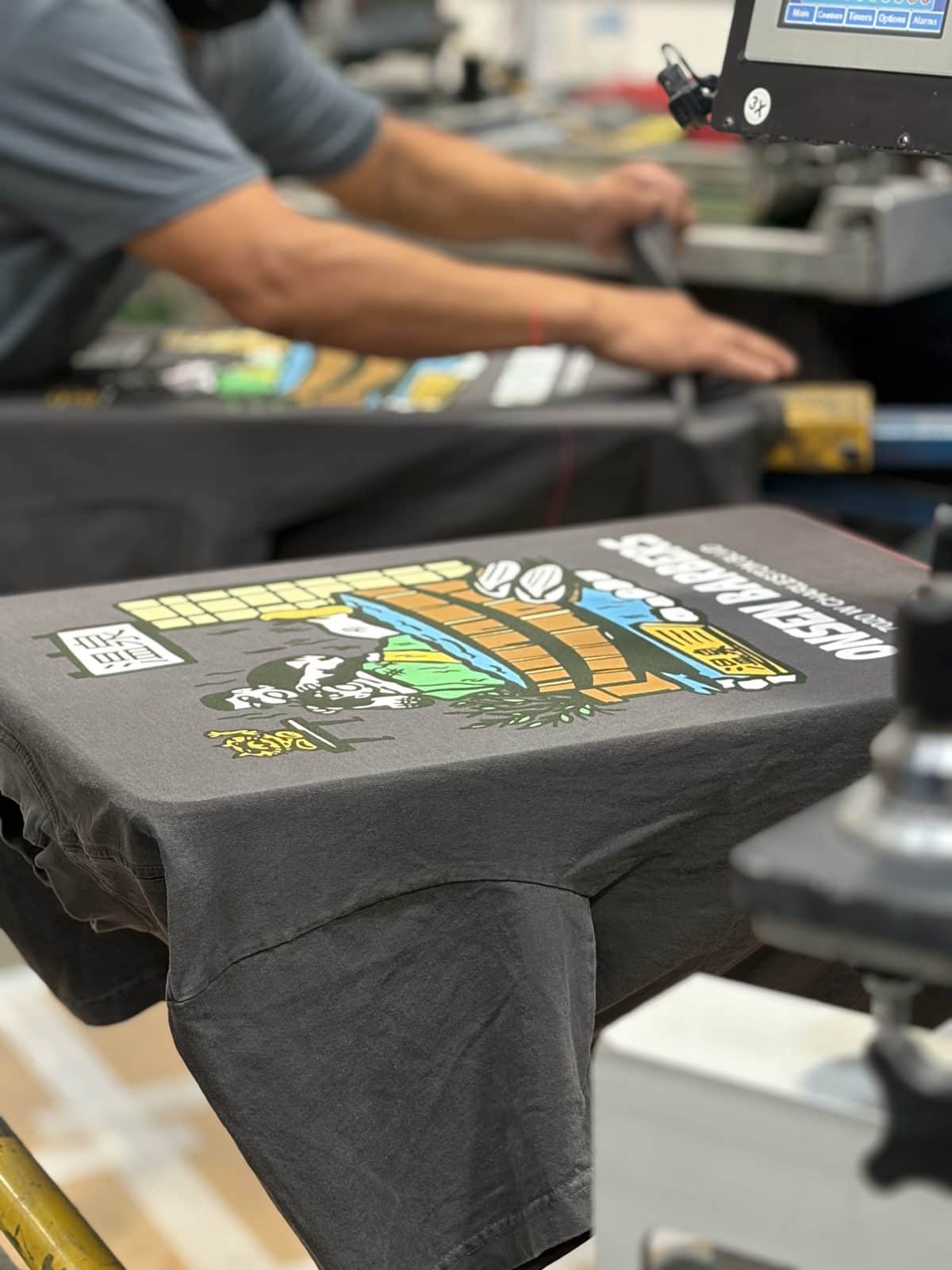 A multi-color design printed on a charcoal t-shirt on the press as a printer works in the background.