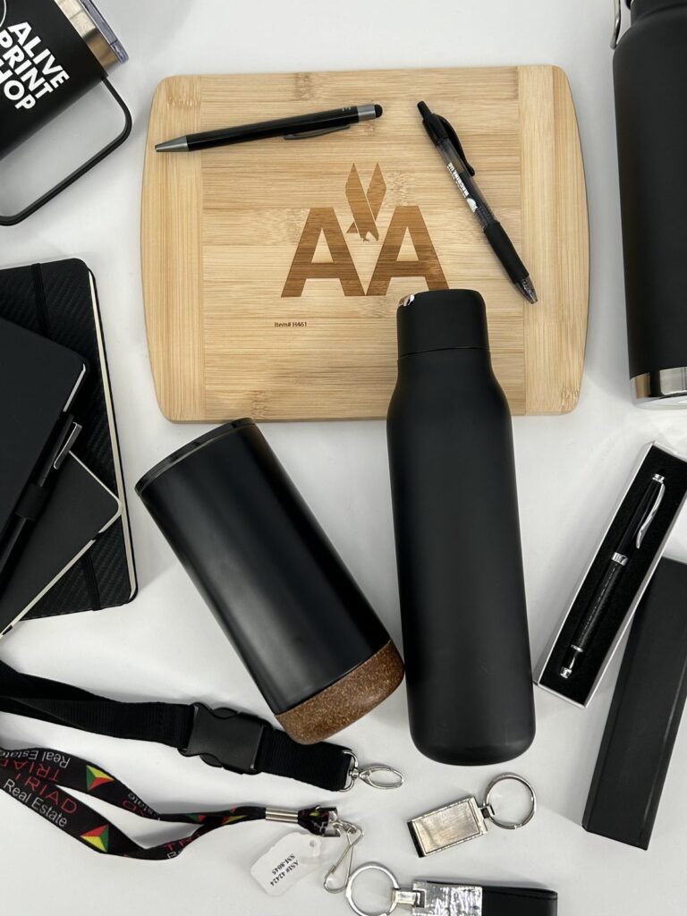 Branded promotional products laid out on a table: an engraved cutting board, pens, insulated bottles, notebooks, lanyards and keychains.